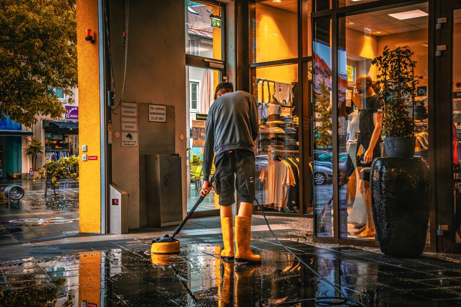A yellow wet floor caution sign with a black and red warning icon is placed on a polished beige tile floor in an indoor space, likely a hallway or entrance area. The sign indicates a potential slip ha