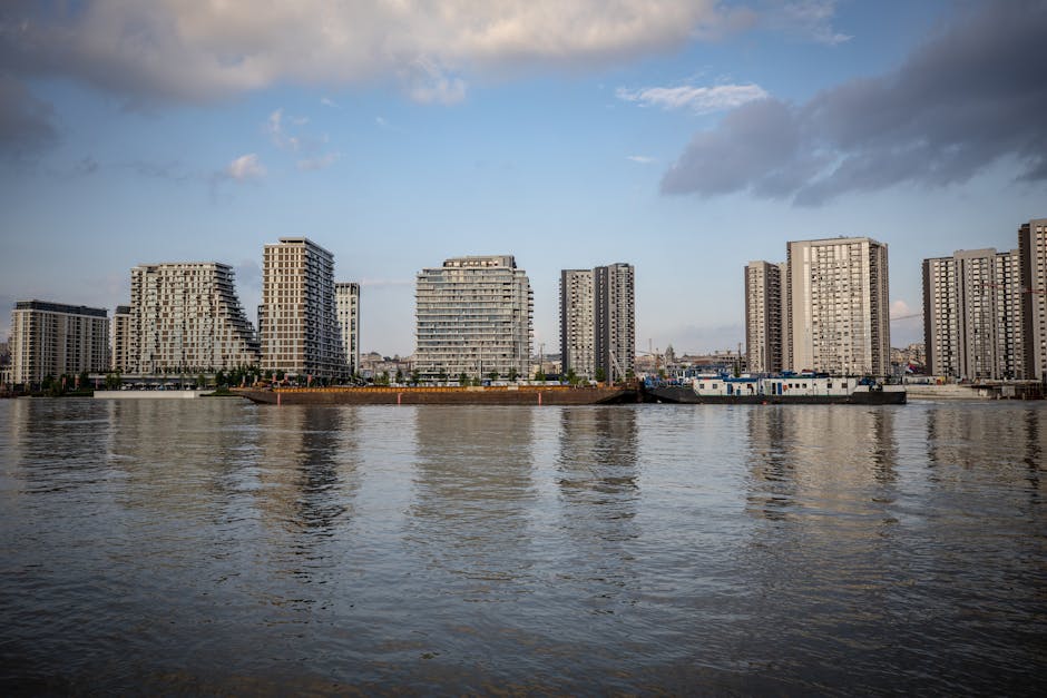 A wide-angle view of a modern city skyline in daylight, featuring tall glass and steel skyscrapers, some with unique architectural designs, set behind a large suspension bridge crossing a river. The b