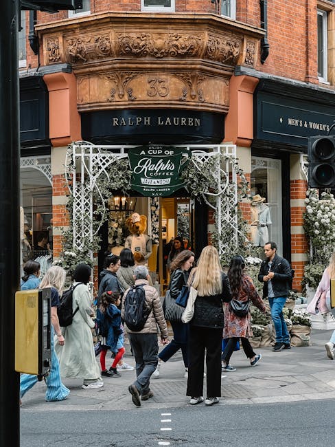 View of the exterior of a Ralph Lauren boutique shop on Kings Road, Chelsea, showing an ornate brick building with detailed architectural embellishments above the entrance, including decorative carvings and the street number '35'. The shop features a black awning with the Ralph Lauren logo and signage, and a white decorative archway framing the entrance. Outside, a group of pedestrians, including men, women, and children, are walking and standing on the sidewalk, some carrying bags or backpacks. The shop window displays mannequins dressed in stylish clothing, with visible flowers and plants adorning the storefront area. The scene is captured in daylight, with natural light illuminating the vibrant street scene and the clean, well-maintained appearance of the shop frontage, reflecting a lively and fashionable urban environment. Carpet Cleaners Chelsea specializes in surface cleaning, deep cleaning, and sanitisation, ensuring hygiene and maintenance for commercial premises like boutique shops.