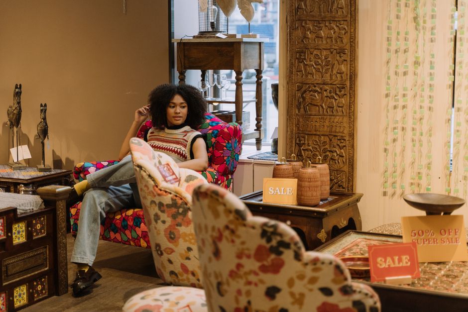 A young woman with curly dark hair is seated comfortably on a vibrant, multicolored upholstered armchair in an interior setting that features wooden and textile decor elements. She is dressed in casual clothing, including a sleeveless striped top and light-colored jeans. The room contains various wooden tables and cabinets displaying decorative items, with some signs advertising sales and discounts, including a yellow 'SALE' sign and a red '50% OFF SUPER SALE' sign. The furniture surfaces, including the plush armchair and tables, appear clean and well-maintained, suggesting regular deep cleaning and surface sanitisation by Carpet Cleaners Chelsea. The lighting is bright, highlighting the clean, dust-free surfaces and the tidy arrangement of the decorative objects, reflecting a space cared for through professional cleaning processes.