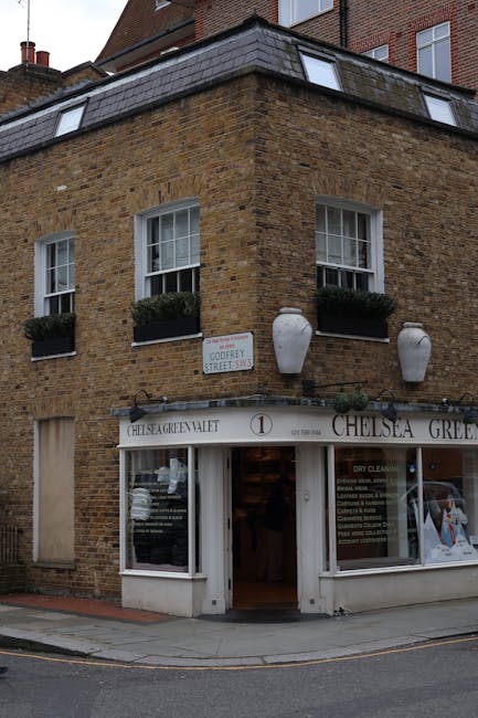 View of the exterior of a Ralph Lauren boutique shop on Kings Road, Chelsea, showing an ornate brick building with detailed architectural embellishments above the entrance, including decorative carvings and the street number '35'. The shop features a black awning with the Ralph Lauren logo and signage, and a white decorative archway framing the entrance. Outside, a group of pedestrians, including men, women, and children, are walking and standing on the sidewalk, some carrying bags or backpacks. The shop window displays mannequins dressed in stylish clothing, with visible flowers and plants adorning the storefront area. The scene is captured in daylight, with natural light illuminating the vibrant street scene and the clean, well-maintained appearance of the shop frontage, reflecting a lively and fashionable urban environment. Carpet Cleaners Chelsea specializes in surface cleaning, deep cleaning, and sanitisation, ensuring hygiene and maintenance for commercial premises like boutique shops.