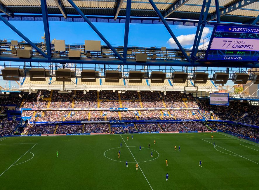 Interior view of a large football stadium with a well-maintained grass pitch. The pitch is marked with white lines for gameplay and has players positioned across the field. The seating stands are fill